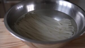 A Stainless Steel Bowl Filled with Water and Soaking Rice Noodles on A Wooden Surface